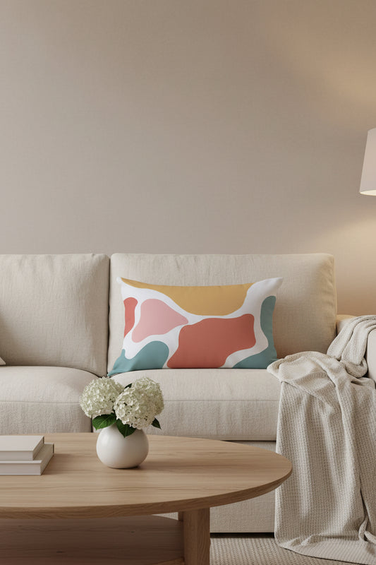 A square throw pillow with an abstract organic pattern in mustard yellow, dusty pink, terracotta red, teal blue, and white, styled on a rustic wooden plank floor next to a white flower arrangement.