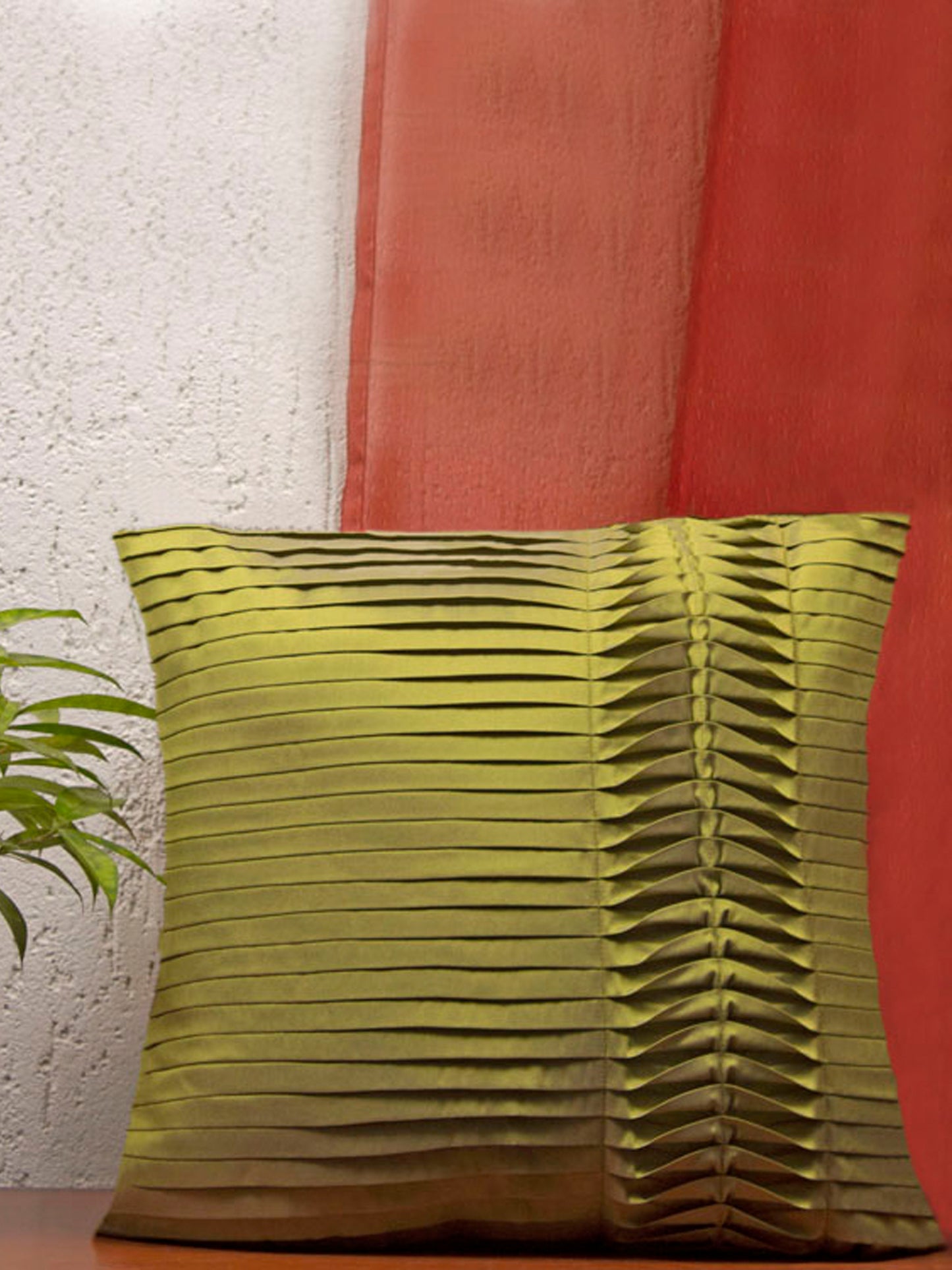 A square olive green throw pillow with a distinctive pleated, accordion-like vertical texture, set against a split background of a white textured wall and a rust-red curtain, with a green plant leaf visible on the left.