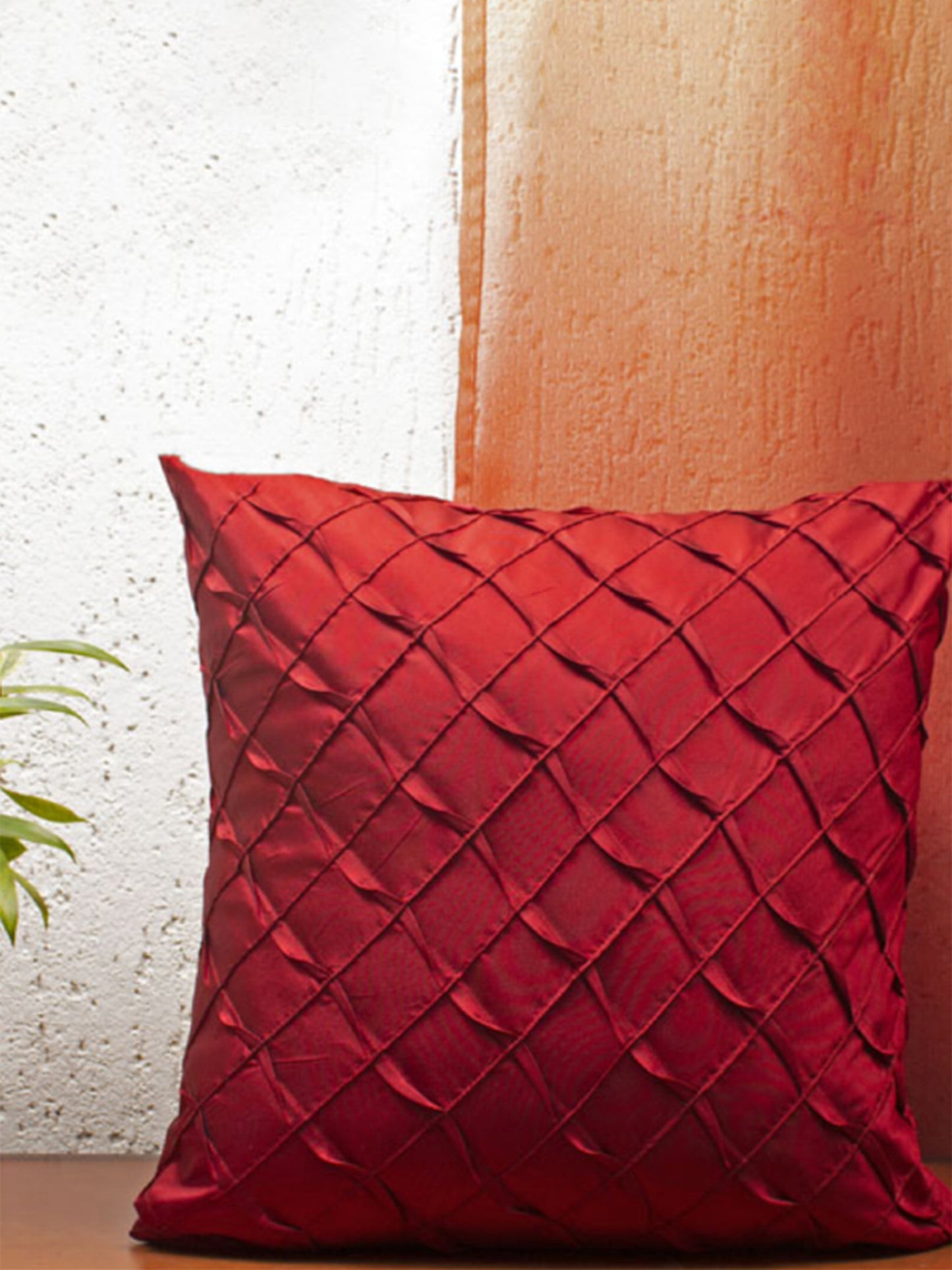 A square red throw pillow with a textured diamond pleated pattern, resting on a wooden surface against a split white and orange textured wall.