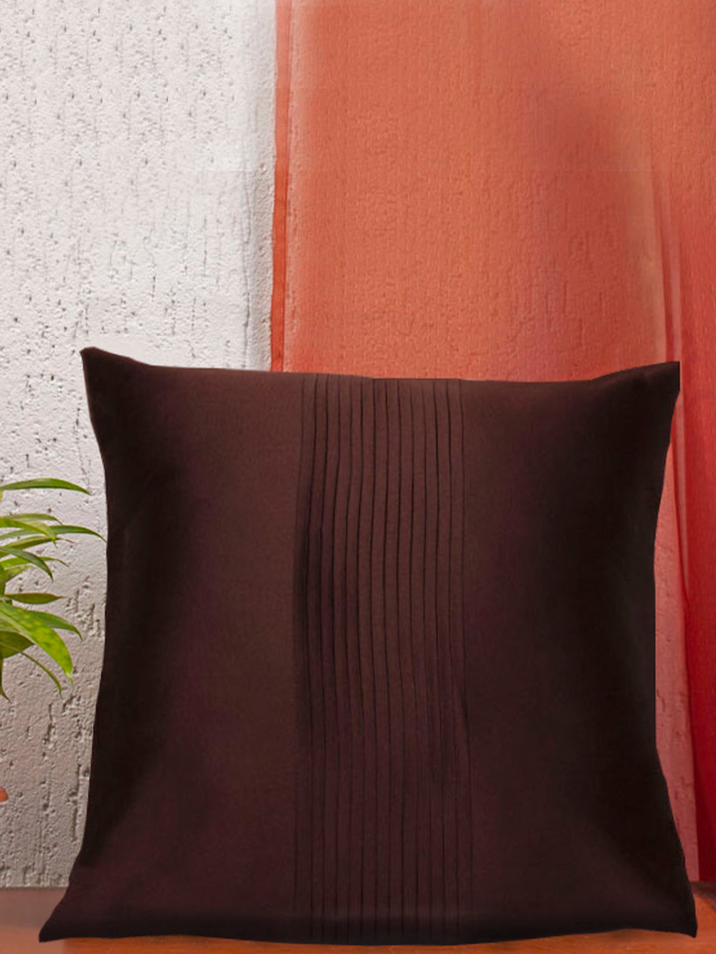 A square dark brown throw pillow with a vertical pleated stripe down the center, set against a split background of textured white and orange walls.