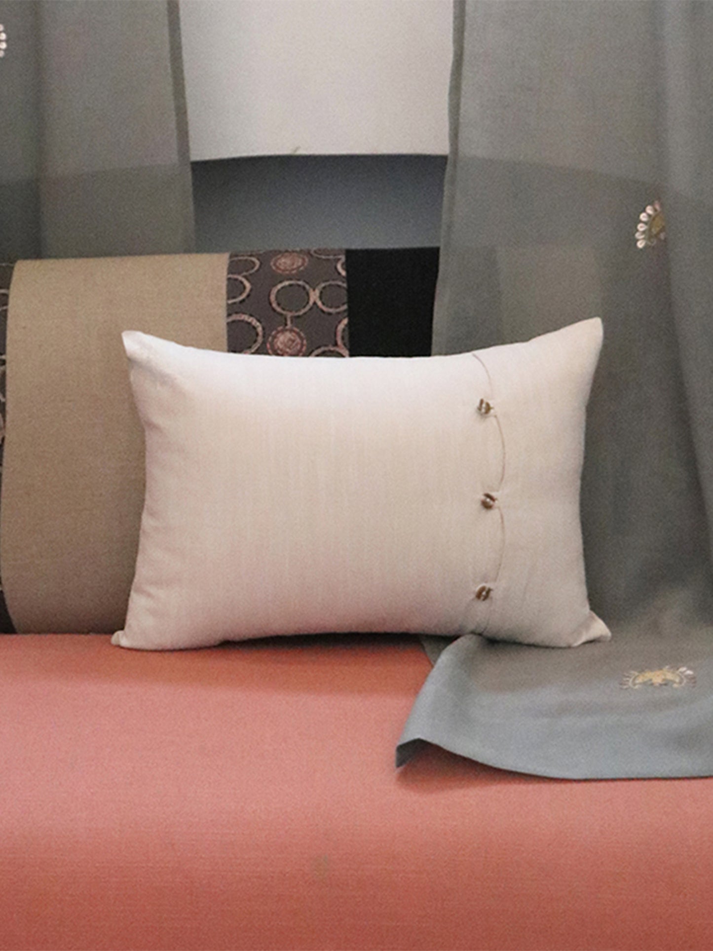 A square off-white throw pillow with three small gold decorative buttons on the right side, resting on a coral-pink sofa in front of a gray and beige patterned backdrop.