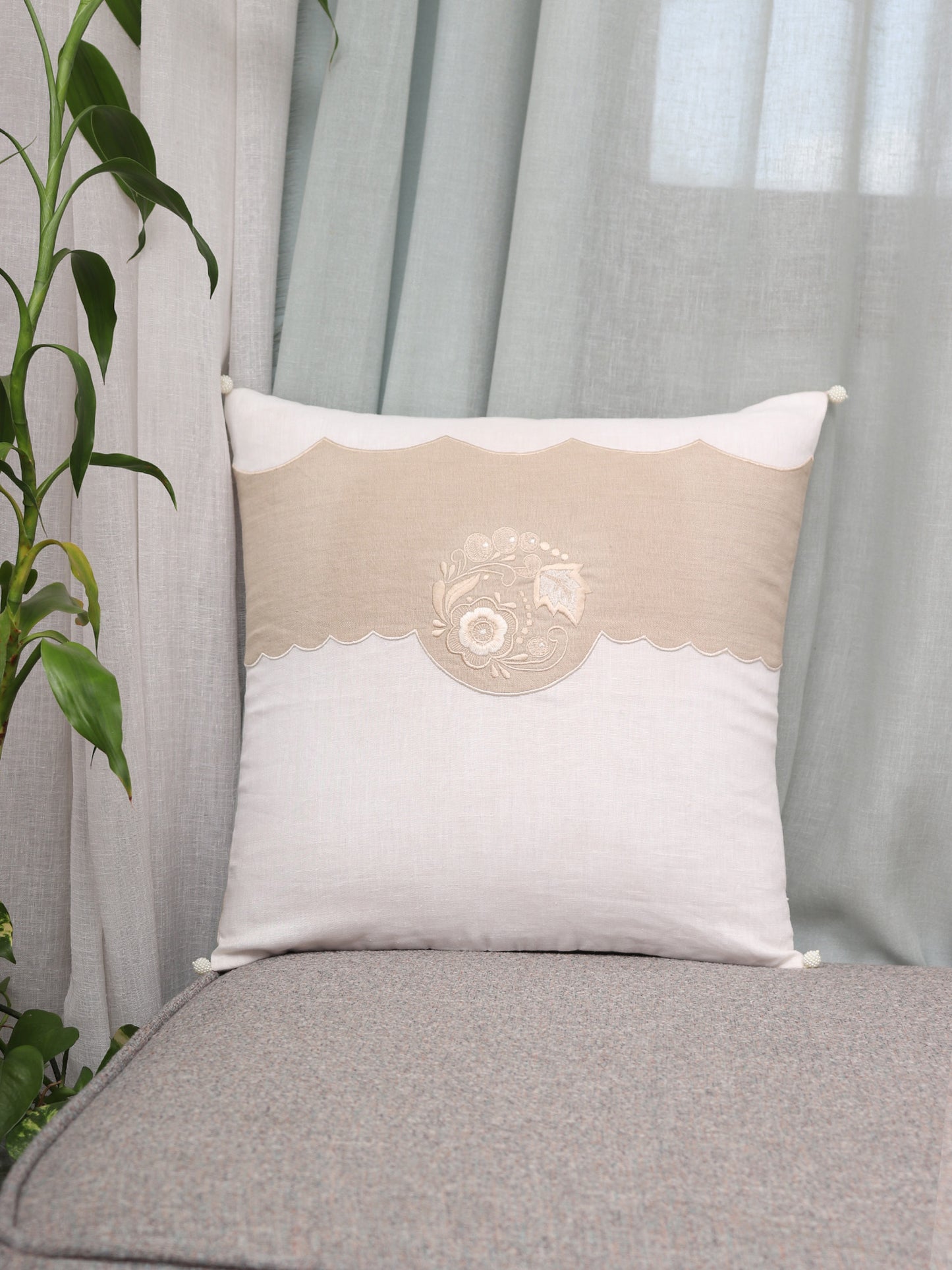A square decorative throw pillow with a beige and off-white design, featuring a central floral embroidery motif and scalloped edges, resting on a gray upholstered bench in front of sheer curtains and next to a green plant.