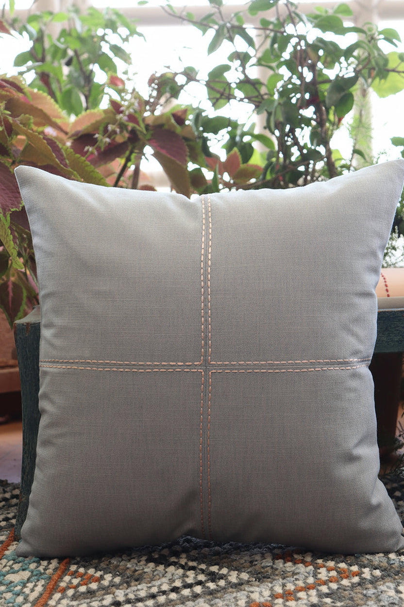 A square gray throw pillow with a four-quadrant stitched design, resting on a patterned rug in front of a wooden bench with green plants in the background.