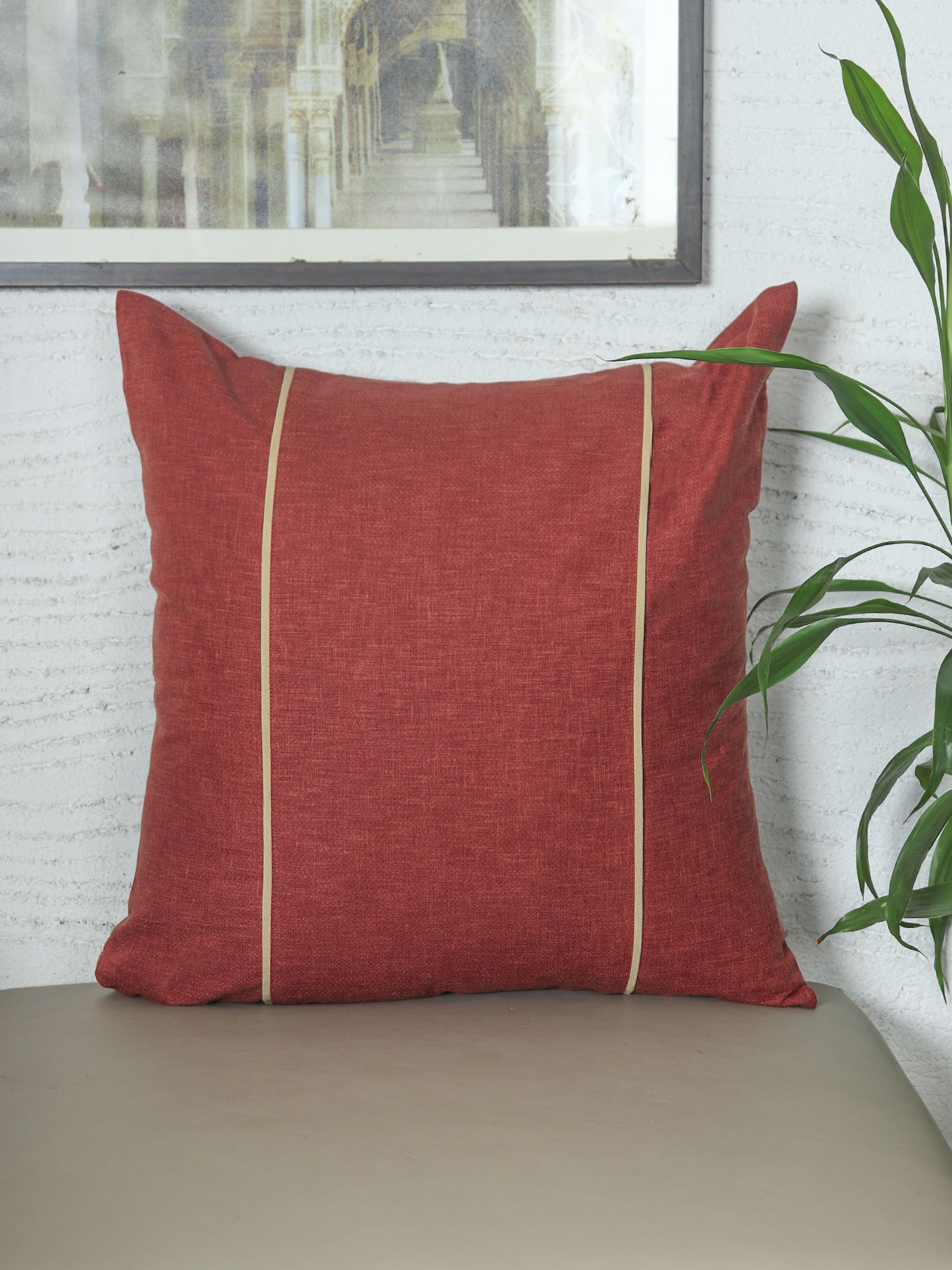 A square terracotta red throw pillow with two vertical cream stripes, resting on a beige surface against a white textured wall with a framed print and green plant leaves in the background.