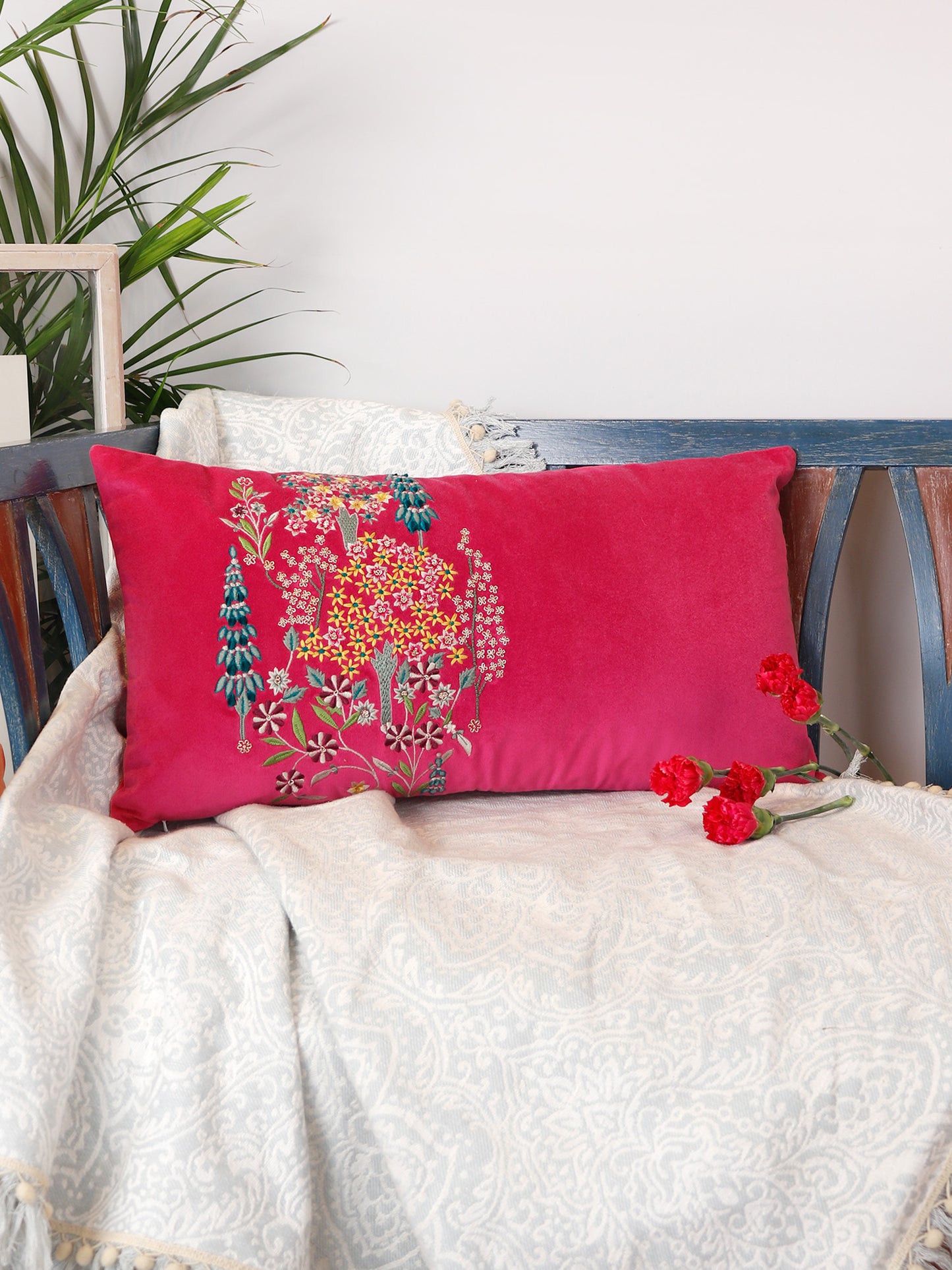 A rectangular pink throw pillow with colorful floral embroidery, resting on a white patterned throw blanket draped over a wooden bench, with red flowers and a green plant in the background.