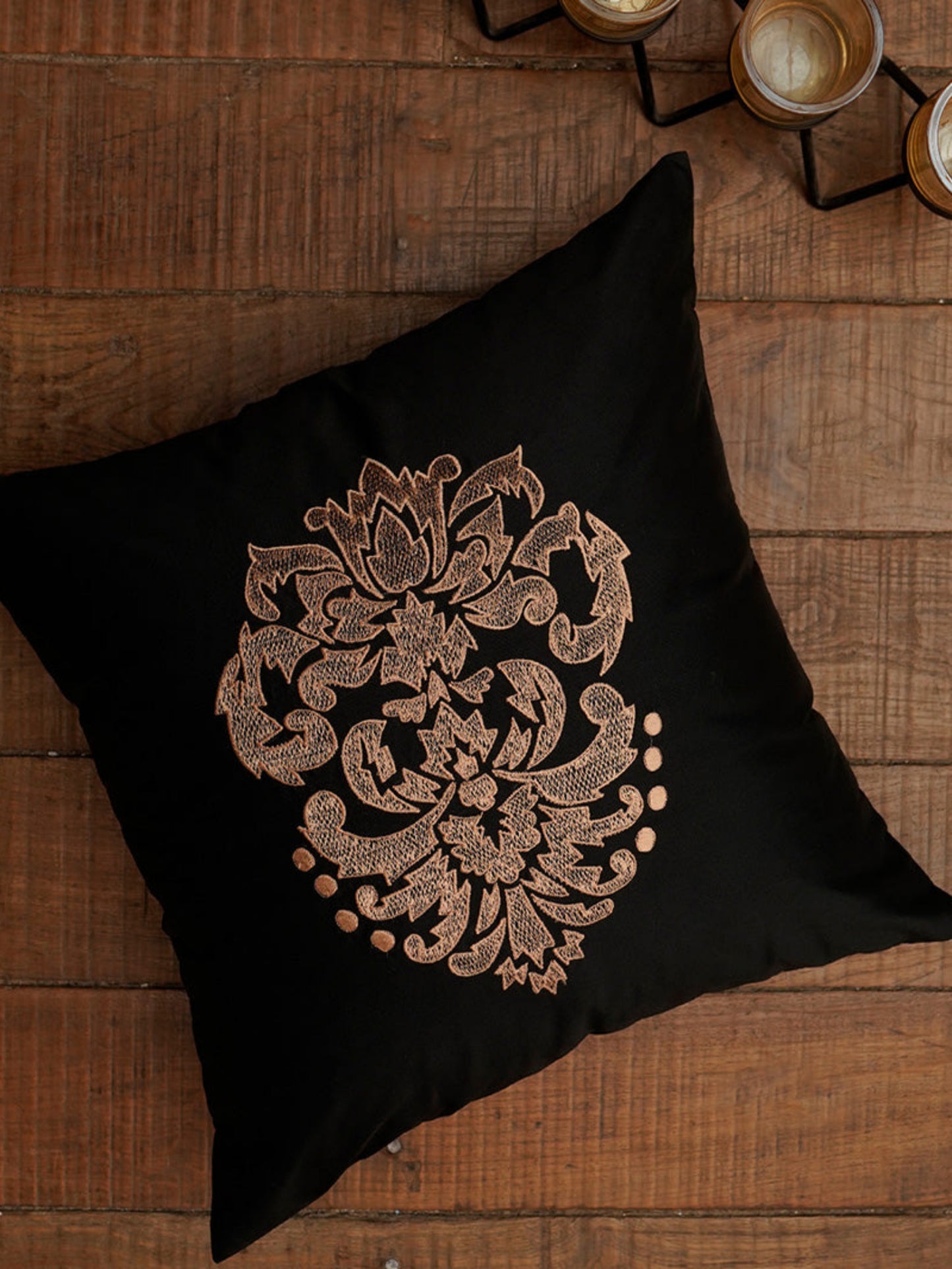A square black throw pillow featuring an intricate metallic rose gold embroidered floral mandala design, displayed on a rustic dark brown wooden surface with decorative candles in the background.