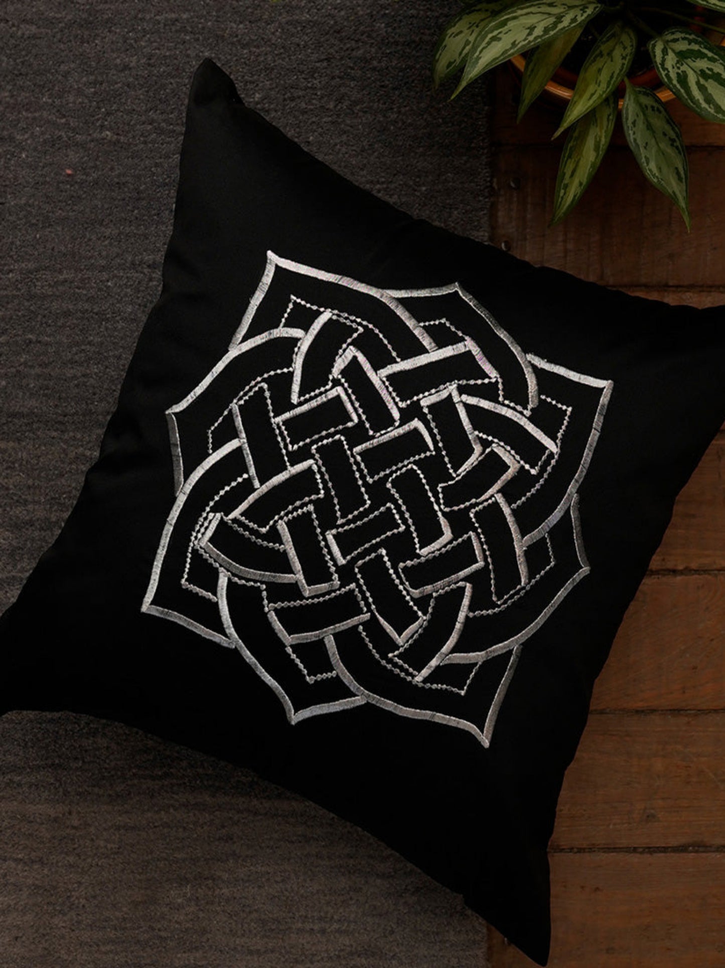 A square black throw pillow featuring a large white Celtic knot design in the center, styled on a dark textured surface next to a wooden floor and green leaves.