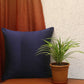 A navy blue square pillow with vertical pleats sits on a wooden bench next to a potted green plant, with a rust-colored curtain and a textured white wall in the background.