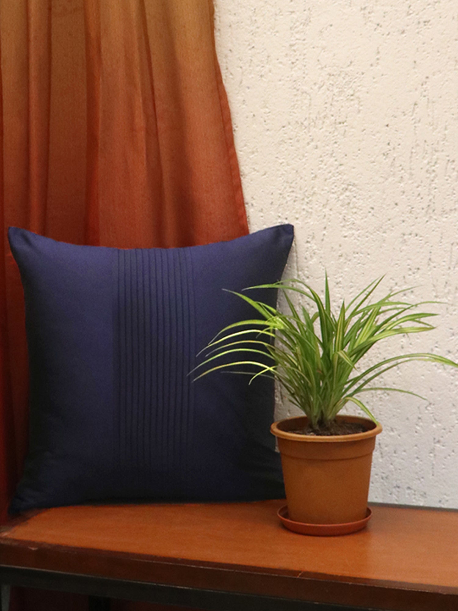 A navy blue square pillow with vertical pleats sits on a wooden bench next to a potted green plant, with a rust-colored curtain and a textured white wall in the background.