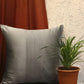 A square gray throw pillow with vertical channel tufting sits on a wooden surface next to a potted green plant, with a rust-colored curtain and textured wall in the background.