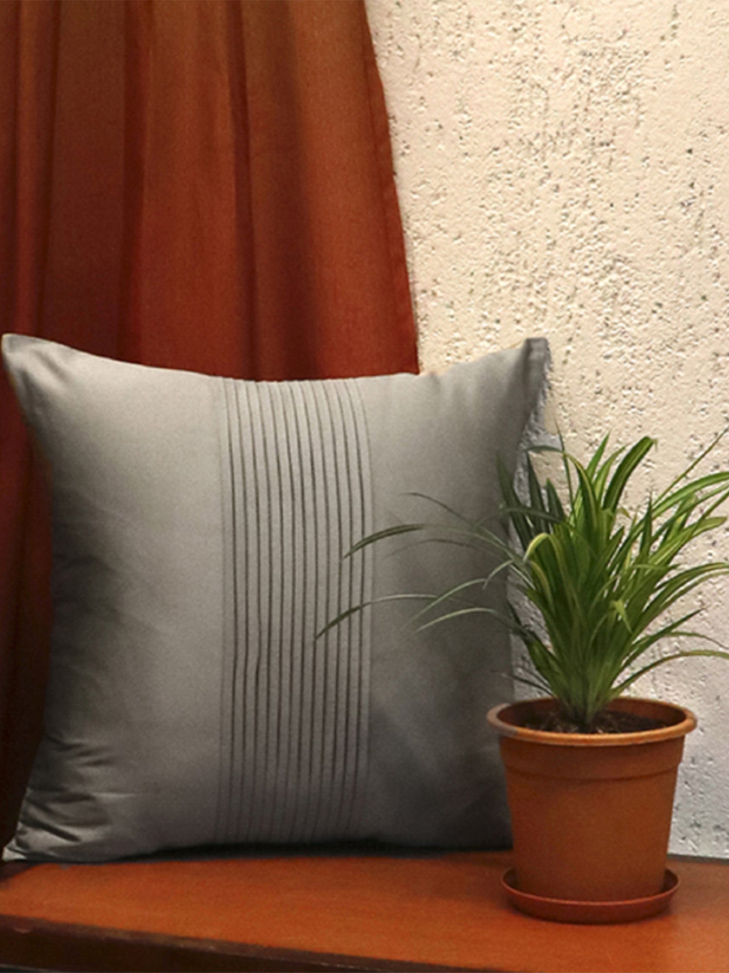 A square gray throw pillow with vertical channel tufting sits on a wooden surface next to a potted green plant, with a rust-colored curtain and textured wall in the background.