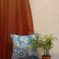 A square decorative throw pillow with a blue and white block-print style pattern, styled on a wooden surface next to a potted green plant and a rust-colored curtain.