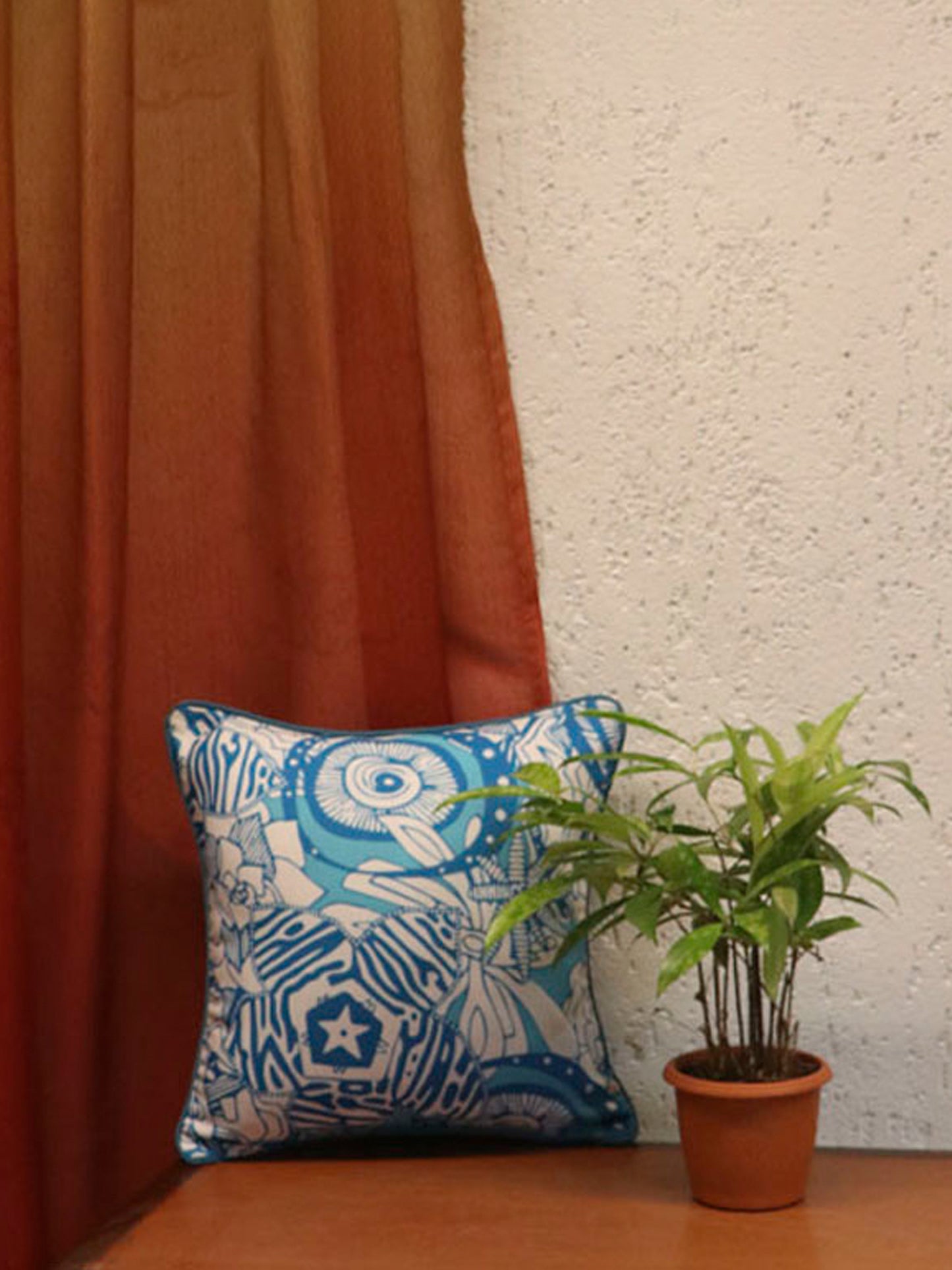 A square decorative throw pillow with a blue and white block-print style pattern, styled on a wooden surface next to a potted green plant and a rust-colored curtain.