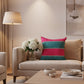 A square decorative throw pillow with horizontal stripes in bright pink and teal, featuring chevron stitching details and gold tassel trim, displayed on a rustic wooden plank background next to a white flower arrangement.