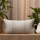 A rectangular off-white throw pillow with three vertical embroidered lines and small blue dots, resting on a beige tablecloth in a styled interior setting with plants and neutral curtains.