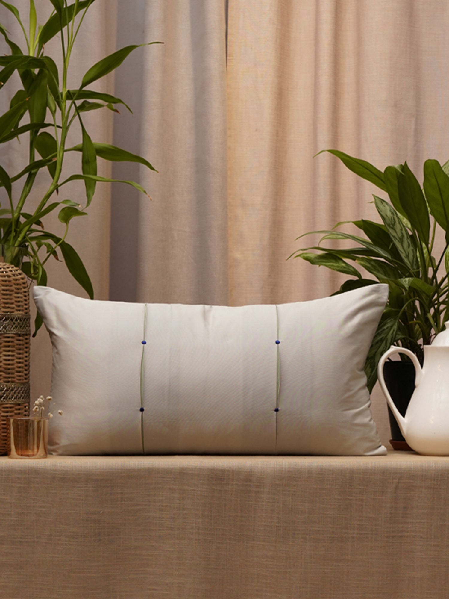 A rectangular off-white throw pillow with three vertical embroidered lines and small blue dots, resting on a beige tablecloth in a styled interior setting with plants and neutral curtains.