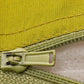 Close-up of a light olive green zipper pull and matching zipper tape against a textured yellow-green fabric background.