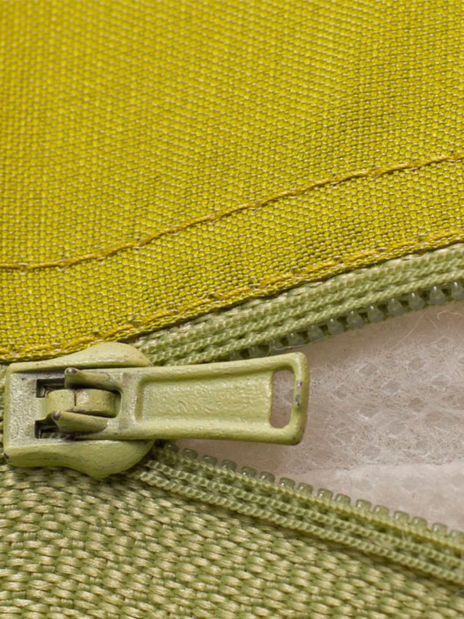 Close-up of a light olive green zipper pull and matching zipper tape against a textured yellow-green fabric background.