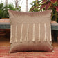 A square decorative throw pillow in a textured taupe-brown fabric, featuring a horizontal band of cream geometric embroidery and five long hanging tassels, resting on a red patterned woven rug with a rustic bench and potted plants in the background.