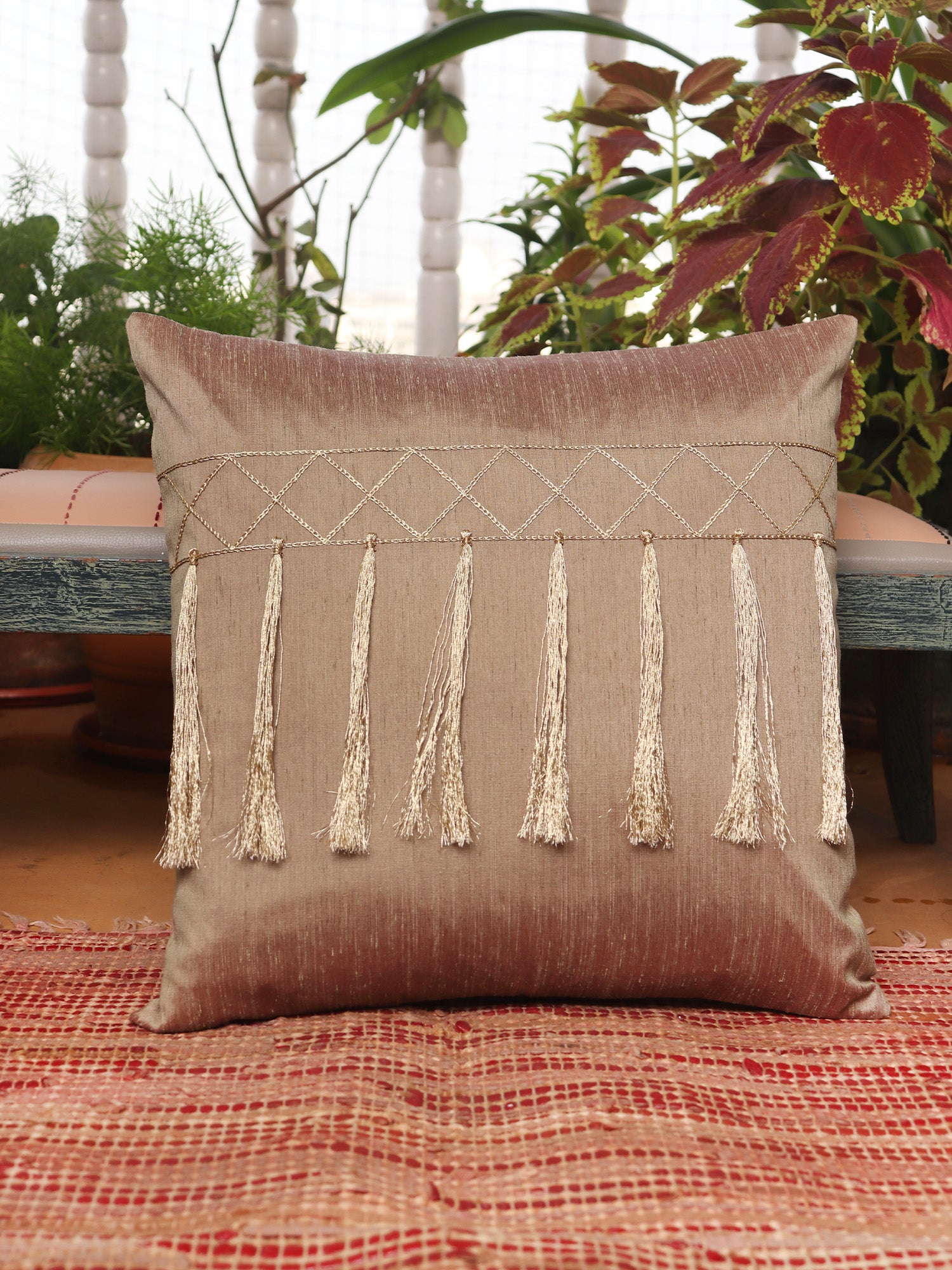 A square decorative throw pillow in a textured taupe-brown fabric, featuring a horizontal band of cream geometric embroidery and five long hanging tassels, resting on a red patterned woven rug with a rustic bench and potted plants in the background.