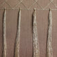 A close-up of a woven textile or wall hanging with a geometric diamond pattern across the top and four long fringe tassels hanging vertically on a brown textured background.