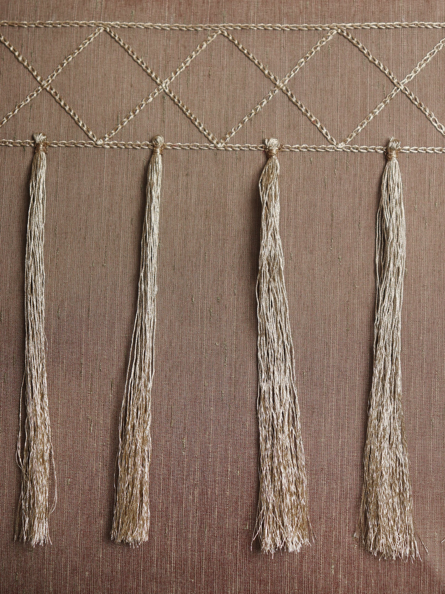 A close-up of a woven textile or wall hanging with a geometric diamond pattern across the top and four long fringe tassels hanging vertically on a brown textured background.