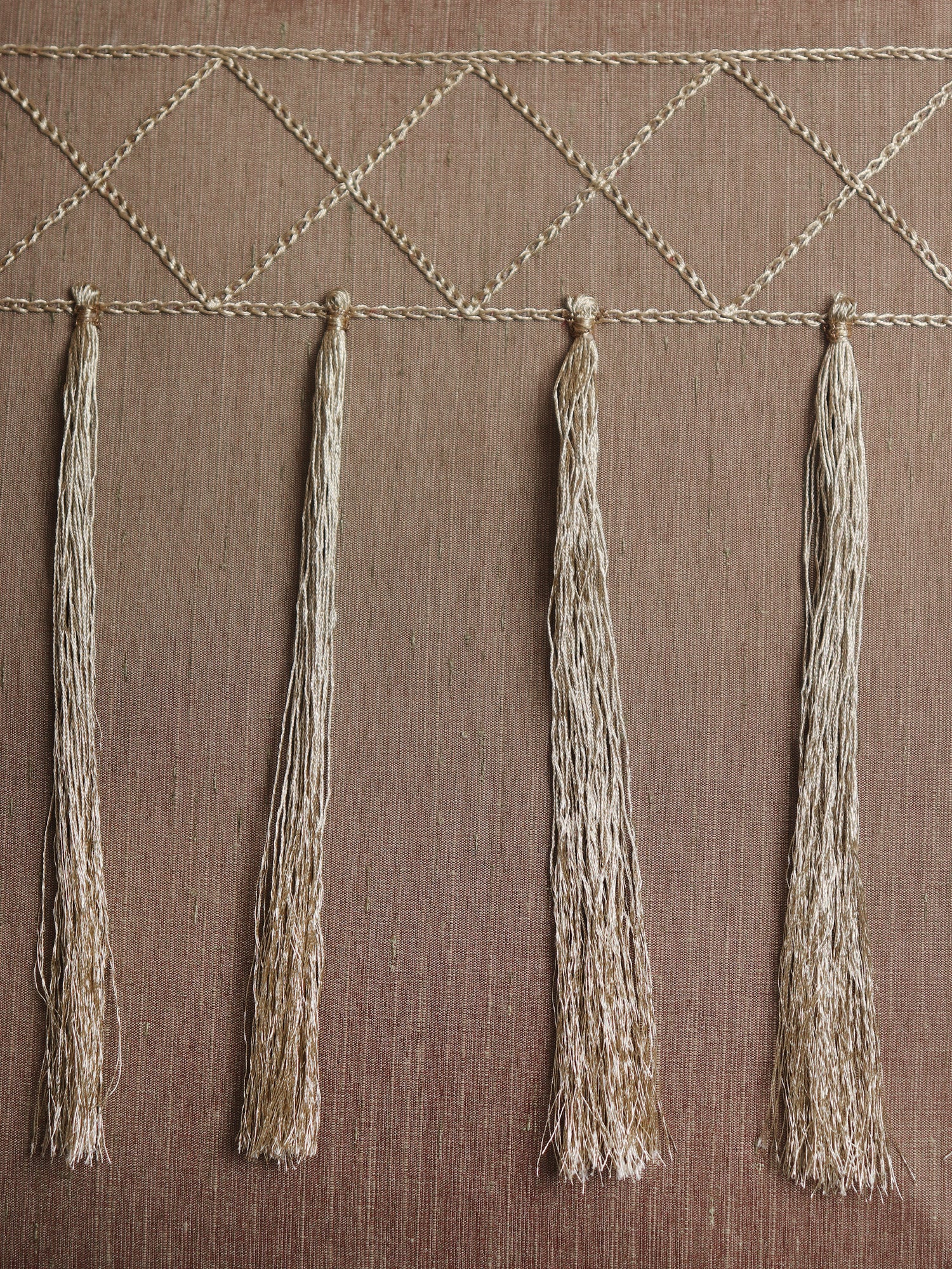 A close-up of a woven textile or wall hanging with a geometric diamond pattern across the top and four long fringe tassels hanging vertically on a brown textured background.