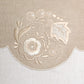 Close-up of a beige embroidered textile detail featuring a scalloped border and floral embroidery in cream and light beige on a woven fabric background.