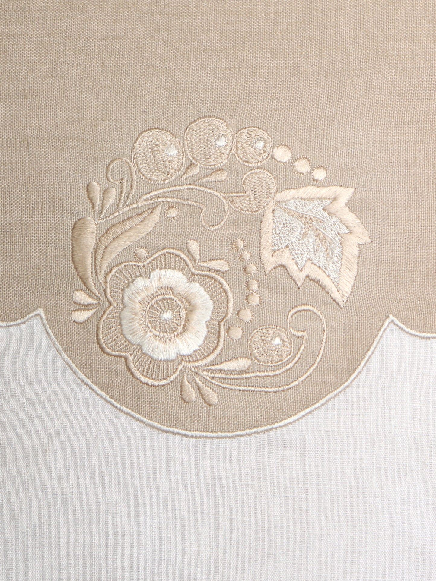 Close-up of a beige embroidered textile detail featuring a scalloped border and floral embroidery in cream and light beige on a woven fabric background.