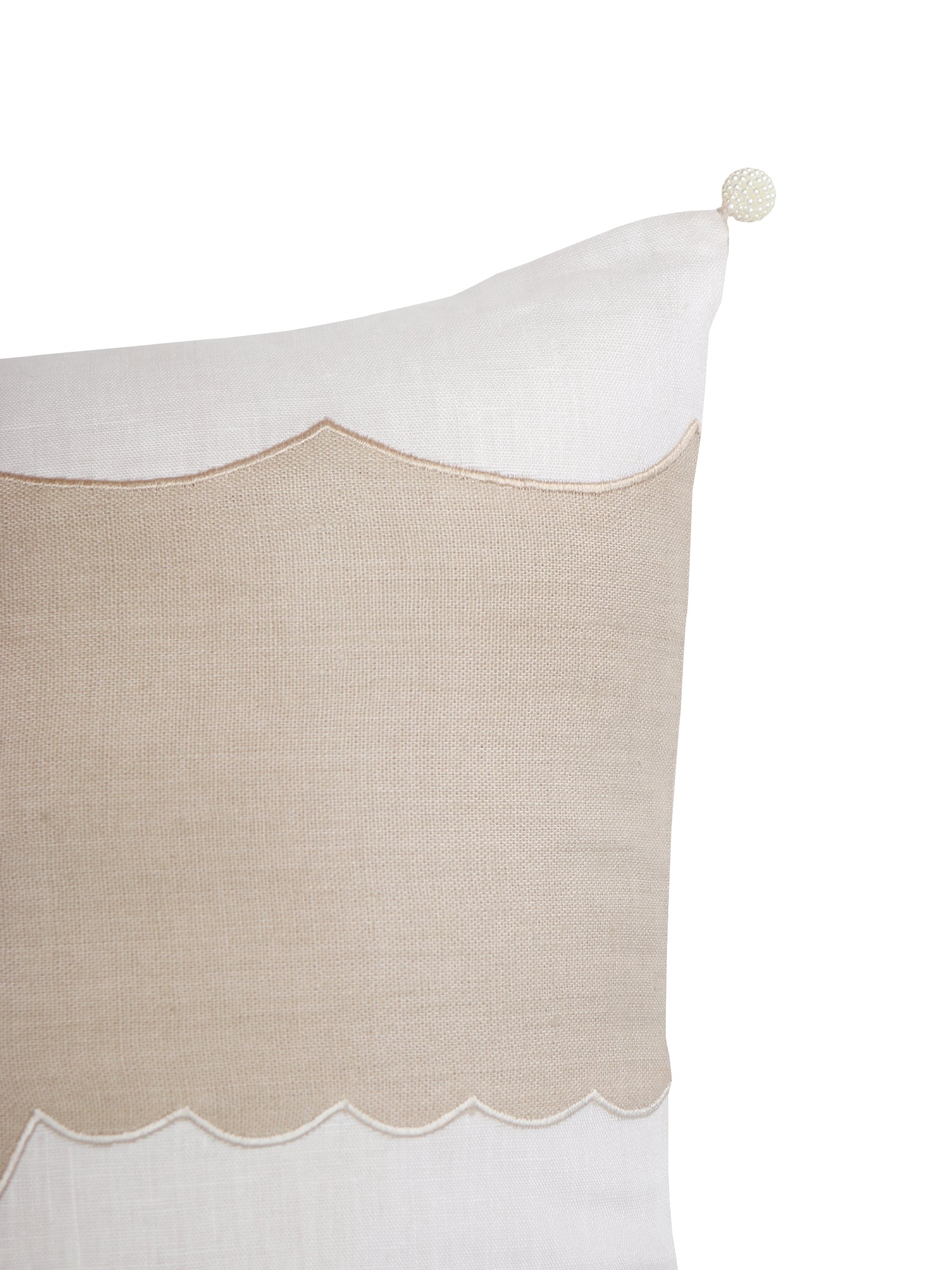 A close-up of a decorative throw pillow featuring a natural beige linen panel with a scalloped edge, bordered by white fabric and finished with a small pom-pom on the corner, set against a plain white background.