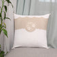 A square decorative throw pillow with a beige and off-white design, featuring a central floral embroidery motif and scalloped edges, resting on a gray upholstered bench in front of sheer curtains and next to a green plant.