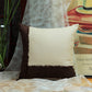 A square decorative throw pillow with a cream center and dark brown fringed borders, styled on a wooden surface with sheer white fabric and a framed print in the background.