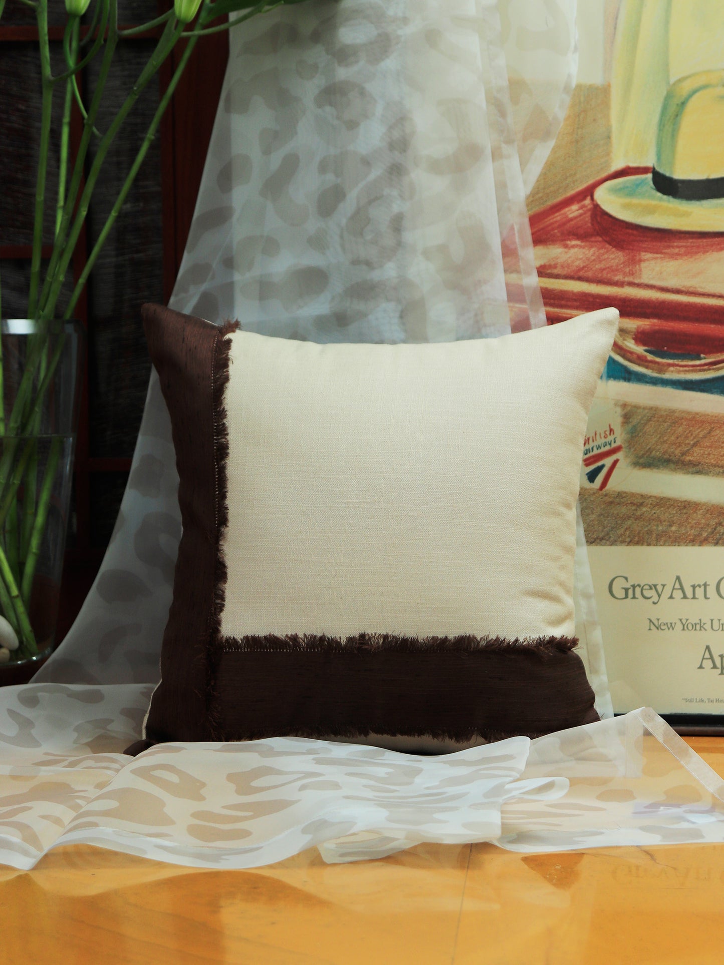 A square decorative throw pillow with a cream center and dark brown fringed borders, styled on a wooden surface with sheer white fabric and a framed print in the background.