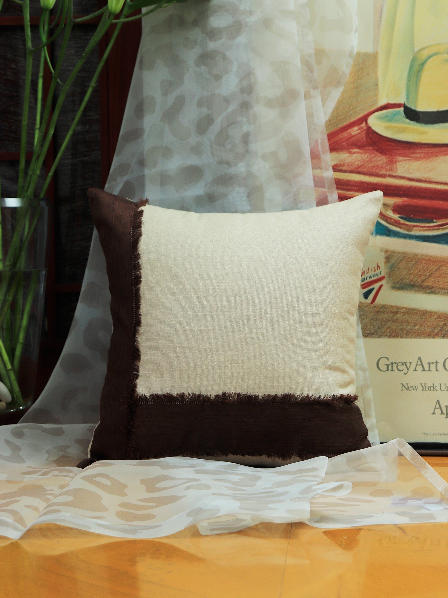 A square decorative throw pillow with a cream center and dark brown fringed borders, styled on a wooden surface with sheer white fabric and a framed print in the background.